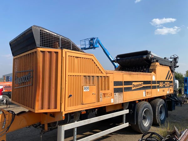 A large orange Doppstadt AK-230 mobile screening/shredding machine on dual axles, parked outdoors. A blue boom lift arm extends over its conveyors, and the machine’s intake hopper and grated vent are visible under a clear blue sky.