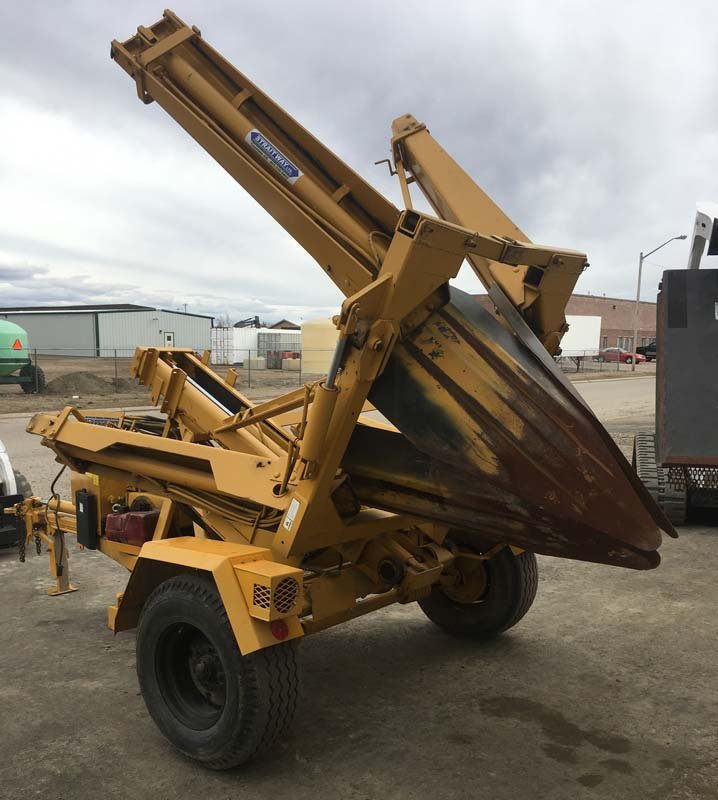 A yellow towable trencher parked on a gravel lot with its dual digging booms raised and folded, showing rusted curved digging blades and black rubber tires under a cloudy sky.