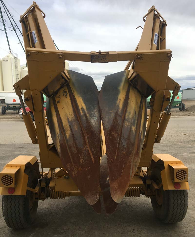 A rear view of a yellow trencher with two large, rusted, curved digging blades folded upright between its wheels under a cloudy sky.