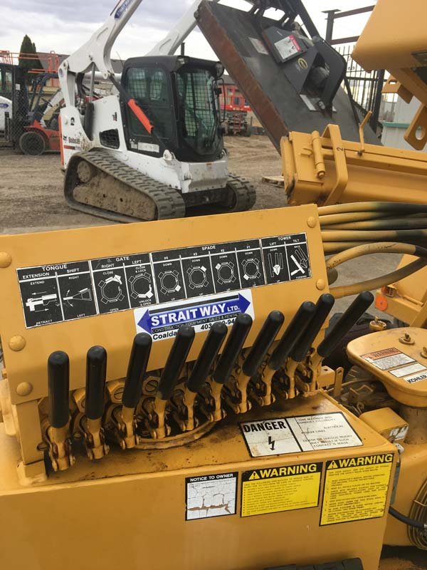 A close-up of a yellow construction machine’s control panel showing multiple black levers, labeled diagrams, and warning stickers. A white skid-steer loader is visible in the background.